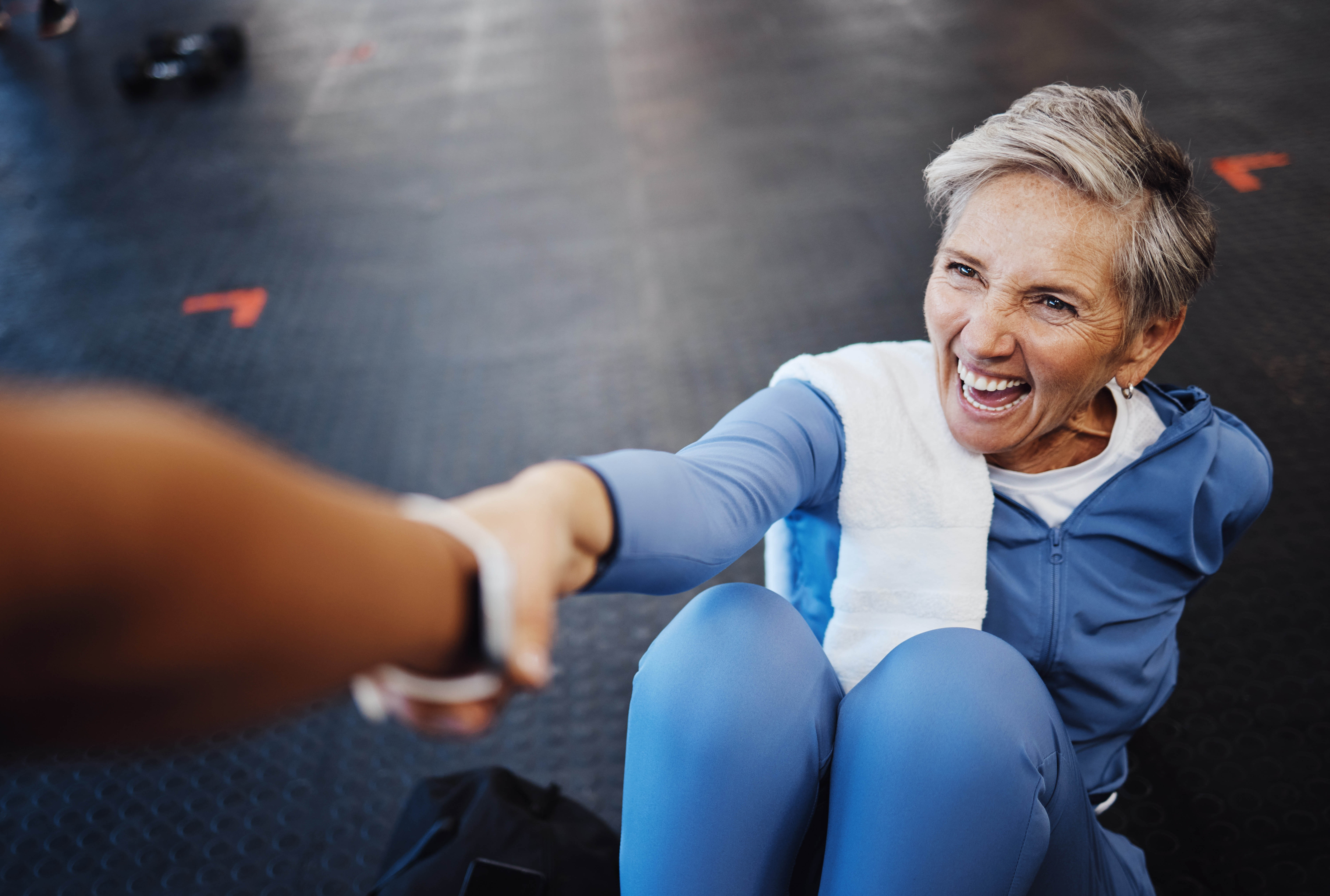Older woman laying on the floor in the gym reaching up towards a hand that is reaching to pull her up Older woman laying on the floor in the gym reaching up towards a hand that is reaching to pull her up